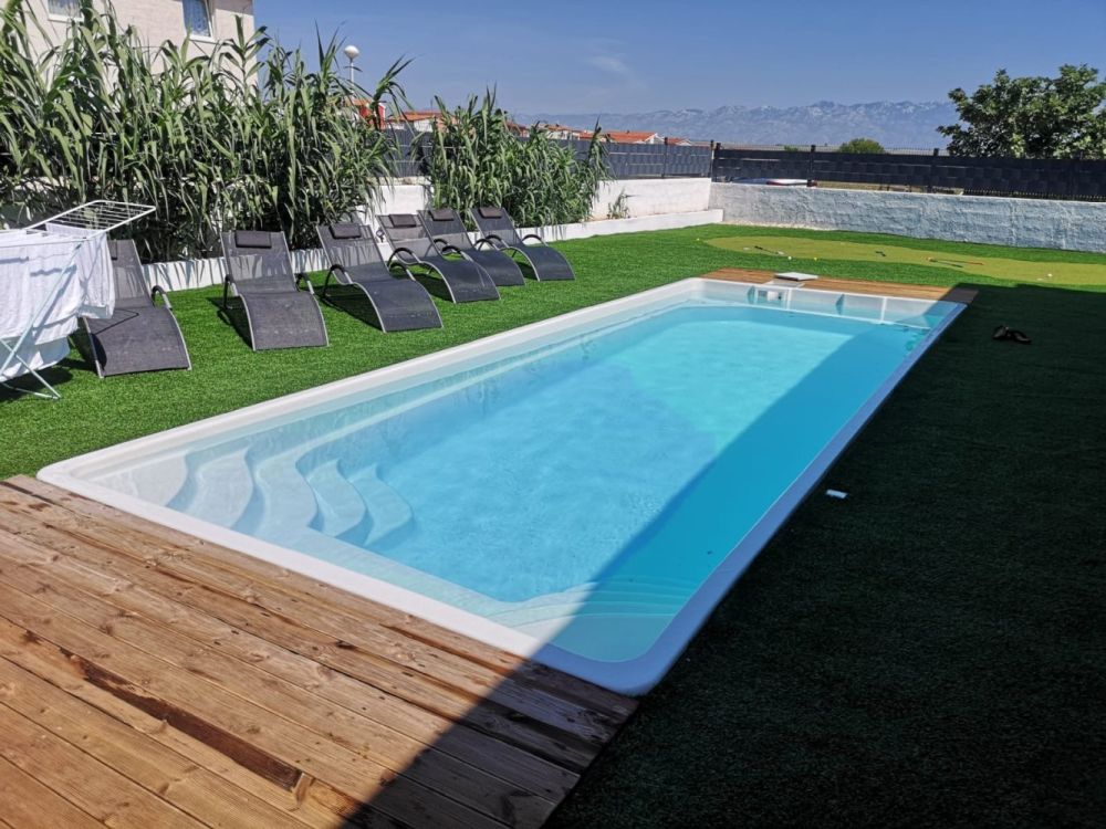 Prostokątny odkryty basen z czystą, błękitną wodą, otoczony sztuczną trawą i drewnianym pomostem. Sześć czarnych leżaków ustawionych jest wzdłuż jednej strony, w pobliżu wysokich zielonych roślin i białego stojaka do suszenia.