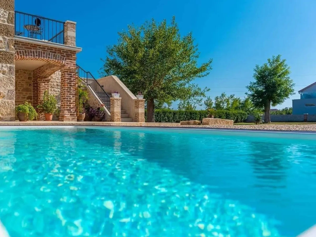 Czysty niebieski basen przed rustykalnym ceglanym budynkiem ze schodami prowadzącymi w górę, otoczony drzewami i jasnym błękitnym niebem w słoneczny dzień.