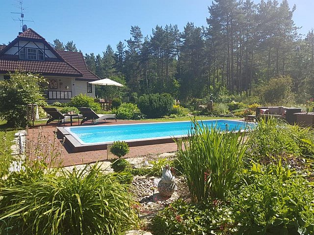 Podwórko z prostokątnym basenem otoczonym zielenią, leżakami i ogrodem. Dom ze spadzistym dachem i drzewa w tle znajdują się pod czystym, błękitnym niebem.