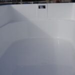 Zbliżenie pustego, białego, plastikowego lub z włókna szklanego kontenera lub skrzyni ładunkowej ciężarówki z wysokimi bokami, widziane od wewnątrz. W rozmytym tle widoczne są pojazdy i budynki.