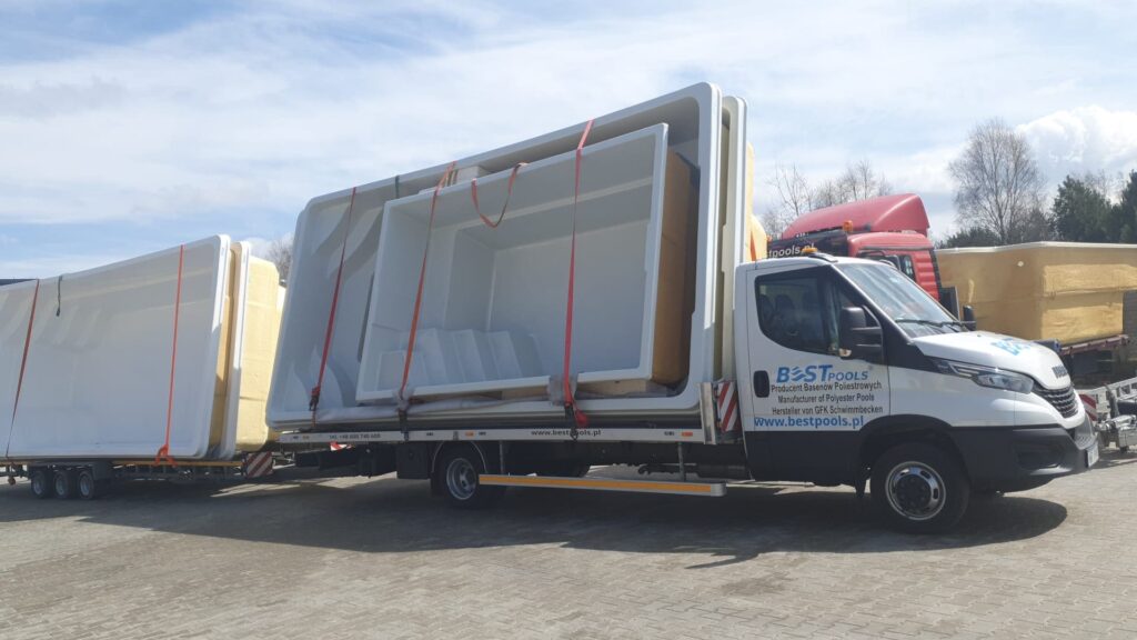 Ciężarówka z platformą załadowaną dużymi, białymi muszlami basenowymi