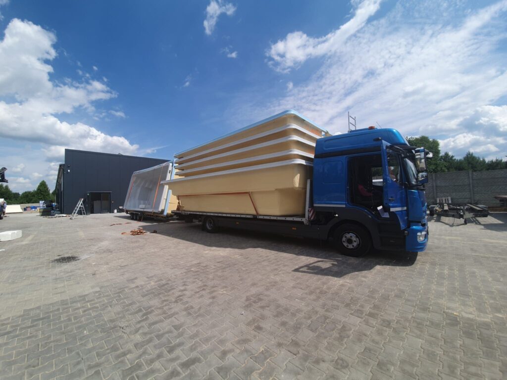 Niebieska ciężarówka z platformą jest zaparkowana na utwardzonej działce, przewożąc duże, ułożone w stosy muszle basenowe