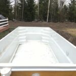 Pusty, prostokątny, biały basen z włókna szklanego jest zainstalowany na podwórku otoczonym ziemią i trawą, z drzewami i białym płotem w tle.