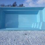 Duża, pusta, jasnoniebieska prostokątna muszla basenowa stoi na zewnątrz na śniegu, z trzema wbudowanymi narożnymi schodkami. W tle widoczne są bezlistne drzewa i częściowo zachmurzone niebo.