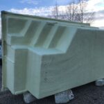Duża, prostokątna, pokryta żółtą pianką konstrukcja stoi na zewnątrz na kamiennych blokach. Powierzchnia ma teksturowane, kanciaste wgłębienia i rowki. W tle znajdują się drzewa, zachmurzone niebo i pojazdy.