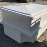 Duży biały plastikowy pojemnik do przechowywania z dopasowaną pokrywą stoi na żwirze na zewnątrz, a światło słoneczne rzuca cienie na jego bok.