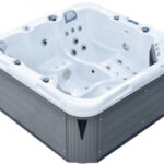 Duża, kwadratowa wanna z hydromasażem na świeżym powietrzu z białym wnętrzem, wieloma wbudowanymi siedzeniami i kilkoma dyszami wodnymi, otoczona szarym panelem zewnętrznym.