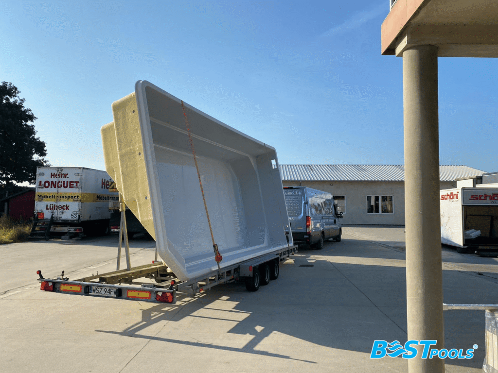 Duży biały basen poliestrowy jest transportowany na przyczepie płaskiej w obszarze przemysłowym, z ciężarówkami i budynkami w tle