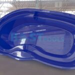 Duża, niebieska muszla basenowa w kształcie nerki jest wyświetlana wewnątrz na wyłożonej kafelkami podłodze z oknami w stylu industrialnym i belkami dachowymi w tle. Basen ma wyprofilowane siedzenia i stopnie.