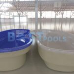 Dwie puste, owalne muszle basenowe są wystawione w pomieszczeniu. Lewy basen jest niebieski, a prawy szary. Oba spoczywają na błyszczącej podłodze wyłożonej kafelkami pod dużym dachem w stylu industrialnym. BEST POOLS jest słabo widoczny w tle.