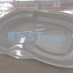 Duży, pusty basen z włókna szklanego o gładkich, zakrzywionych krawędziach znajduje się wewnątrz na wyłożonej kafelkami podłodze pod metalowo-szklanym dachem. Napis BEST POOLS jest słabo widoczny na powierzchni basenu.