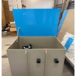 Duży prostokątny pojemnik z dwoma czarnymi łącznikami rur i częściowo otwartą niebieską pokrywą, umieszczony w obszarze przemysłowym lub magazynowym z pudełkami i materiałami w tle.