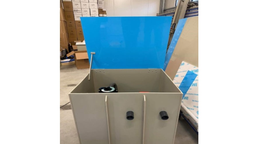 Duży prostokątny pojemnik z dwoma czarnymi łącznikami rur i częściowo otwartą niebieską pokrywą, umieszczony w obszarze przemysłowym lub magazynowym z pudełkami i materiałami w tle.